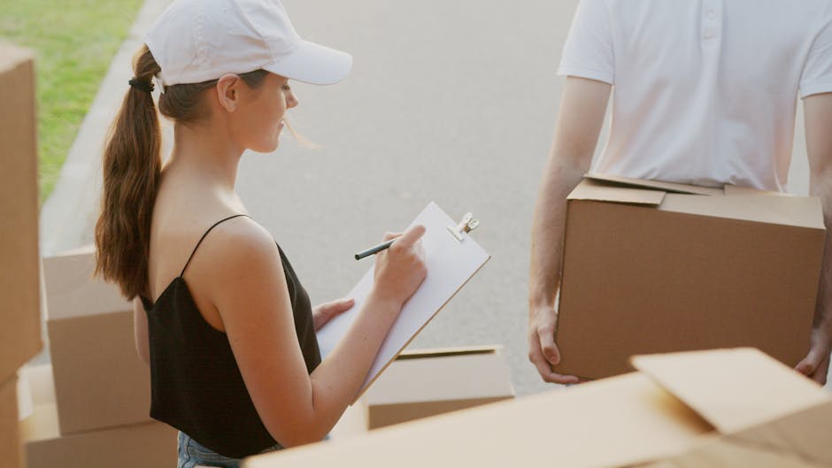 A woman wearing a white cap and black sleeveless top is holding a clipboard with a pen, taking notes during a home relocation or packing process. She stands next to a man dressed in a white t-shirt, who is carrying a medium-sized cardboard box. They are outdoors on a paved surface near the entrance of a property, with several packing boxes stacked nearby. The woman is positioned to the left, with her profile visible, while the man is on the right, partially out of frame. The scene is illuminated by natural daylight, emphasizing the organized packing and loading activities typical of furniture transport or removals. The setting suggests an active moving or packing operation, with the presence of cardboard boxes, packing materials, and the emphasis on loading and documentation as part of a professional removals service by Man with Van Woolwich, supporting the context of house moves and logistics.