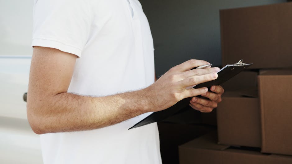 A woman wearing a white cap and black sleeveless top is holding a clipboard with a pen, taking notes during a home relocation or packing process. She stands next to a man dressed in a white t-shirt, who is carrying a medium-sized cardboard box. They are outdoors on a paved surface near the entrance of a property, with several packing boxes stacked nearby. The woman is positioned to the left, with her profile visible, while the man is on the right, partially out of frame. The scene is illuminated by natural daylight, emphasizing the organized packing and loading activities typical of furniture transport or removals. The setting suggests an active moving or packing operation, with the presence of cardboard boxes, packing materials, and the emphasis on loading and documentation as part of a professional removals service by Man with Van Woolwich, supporting the context of house moves and logistics.