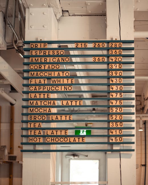 A menu board mounted on a wall inside a cafe or coffee shop displays various beverage options and prices, with each item written on individual orange-colored strips attached to horizontal blue supports. The menu includes drinks such as drip, espresso, americano, cortado, macchiato, flat white, cappuccino, latte, matcha latte, mocha, and various types of tea and hot chocolate, with prices ranging from £215 to £450. Below the menu, a green emergency exit sign is visible, indicating the direction to an exit. The location appears to have an industrial or modern decor style, with an illuminated space and a doorway seen at the bottom of the image, providing natural light to the interior. The setup suggests a functional and organized environment typical for a coffee shop that may involve mobile or flexible seating arrangements and efficient customer service, aligning with the services provided by professional removal and transport specialists like Man with Van Woolwich.