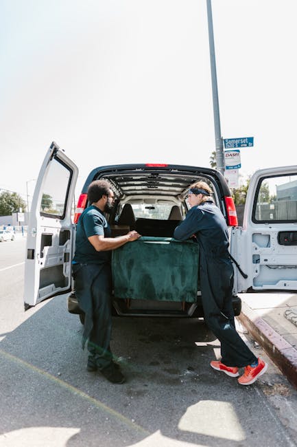 Two movers from Man with Van Woolwich are engaged in a home relocation task, carefully loading a large, rectangular piece of furniture wrapped in dark green protective fabric into the spacious rear cargo area of a white van. The van's rear doors are wide open, revealing the vehicle's interior with additional packing materials and other household items visible inside. The movers are standing on a paved area next to a curb on a partly sunny day, with a street sign and a lamppost visible in the background. This loading process is part of furniture transport and packing and moving services, with the team ensuring secure and professional handling of bulky items for a property relocation in the SE18 postcode area.