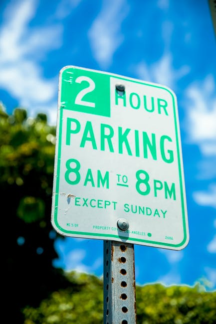 Close-up image of a green and white parking restriction sign affixed to a metal pole against a bright blue sky with some clouds. The sign indicates a two-hour parking limit from 8 am to 8 pm, Monday to Saturday, with an exception on Sundays. In the background, blurred green foliage and the top of a building are visible. This parking sign is relevant to house removals and home relocation services, as it provides information about parking regulations in Woolwich, which Man with Van Woolwich may need to consider when planning furniture transport or loading processes for local moves.