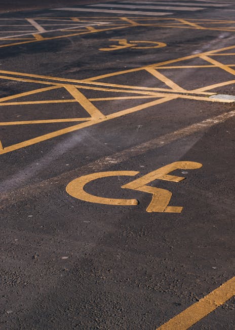 The image shows an outdoor parking area with multiple yellow lines and parking bay markings on a dark asphalt surface. Prominently visible are two designated accessible parking spaces, each marked with a yellow wheelchair symbol and delineated by yellow grid lines, indicating spaces reserved for disabled parking. The surface appears clean and well-maintained, with clear, freshly painted markings. The surrounding environment is unspecified, but the markings suggest a commercial or residential parking lot used for vehicle access to nearby buildings, possibly including a home or business location involved in furniture transport or relocation services. This setting relates to the logistics of home relocation, with the accessibility markings highlighting the importance of designated parking permits, such as those issued by Greenwich Council for Woolwich movers, as referenced on the page for parking permits for moving services.