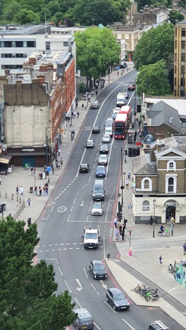 A street scene on Plumstead Road in Woolwich during daylight, featuring parked and moving vehicles including a silver Audi car parked on the right side of the road, a red double-decker bus approaching from the distance, and several other cars along the street. The pavement on both sides is lined with street poles, signposts indicating parking restrictions and a speed limit of 20 miles per hour, and lampposts. On the left, there are residential buildings with brick facades, some with small front gardens, and trees with budding leaves indicating early spring. The street appears to be part of a home relocation or furniture transport process, with visible activity around local buildings. The background shows a mix of urban infrastructure, including more buildings and distant cityscape features under an overcast sky, suggesting a typical urban neighbourhood in Woolwich where Man with Van Woolwich offers removals and moving services.