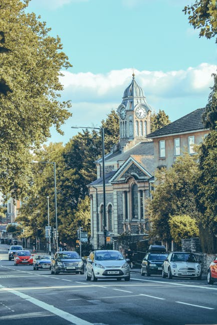 A street scene in Woolwich featuring several parked cars along the roadside with a large historic building in the background, showcasing a tall clock tower with a pointed roof and a clock face on each side. The building appears to be a church or civic structure with brick and stone details. Surrounding the scene are mature trees with green foliage, and a clear blue sky with some scattered clouds. This image captures a typical urban environment suitable for house removals and furniture transport, as part of home relocation services provided by Man with Van Woolwich. The street’s wide pavement and marked lanes suggest an area where loading and unloading furniture or boxes might take place during a relocation process, with natural light highlighting the exterior architecture and nearby vehicles, aligning with the theme of moving logistics and packing and moving operations.