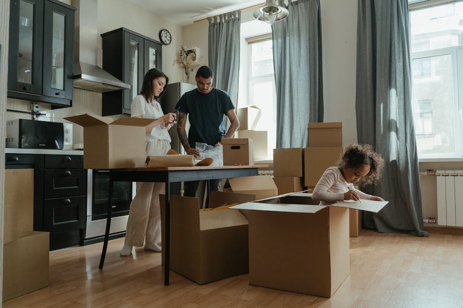 Inside a well-lit room with large windows covered by grey curtains, two adults and a young girl are engaged in packing or unpacking as part of a home relocation process. The woman on the left, dressed in light-colored clothing, is holding a smartphone, while the man in the center, wearing a dark t-shirt, is looking at an object in his hands, possibly assembling or inspecting packing materials. The young girl on the right, with curly hair, is focused on a clear plastic container or tray placed inside a large cardboard box on the wooden floor. The room contains several cardboard moving boxes of various sizes, some open and some sealed, with packing materials like paper or bubble wrap visible next to them. Items such as a microwave, kitchen cabinets, and a wall clock are seen in the background, indicating a kitchen area, likely in the process of packing belongings for a house move. The scene captures the typical logistics involved in household packing and furniture transport, aligning with the services offered by Man with Van Woolwich as part of their removals services, supporting a family’s move near Woolwich Common.