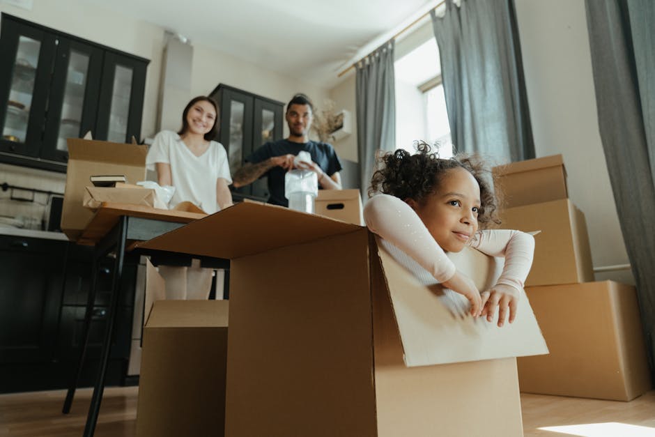 Inside a well-lit living room with large windows and light-colored curtains, a young girl with curly hair is leaning over the edge of a cardboard box, resting her arms on the box's flaps, looking out with a slight smile. Behind her, two adults, a woman with shoulder-length brown hair and a man with short dark hair and a beard, are standing near a table covered with unpacked moving boxes and packing materials, such as brown cardboard boxes, some open with packing paper and plastic wrapping visible. The room features dark-colored cabinets and a modern, minimalistic decor. The scene captures the process of home relocation, with furniture and household items being prepared for transport, as part of a furniture transport or packing and moving operation carried out by a professional removals service, such as Man with Van Woolwich, supporting families moving near Woolwich Common.
