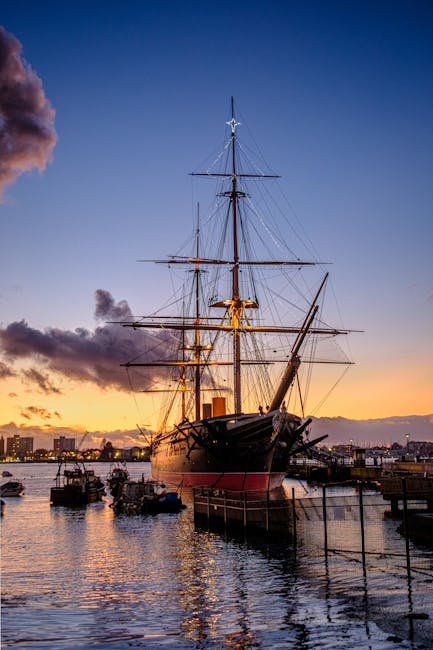 A large historic sailing ship with multiple masts and rigging is moored at a dock during sunset, with the sky displaying orange, pink, and purple hues. The vessel's hull is painted black with red acce