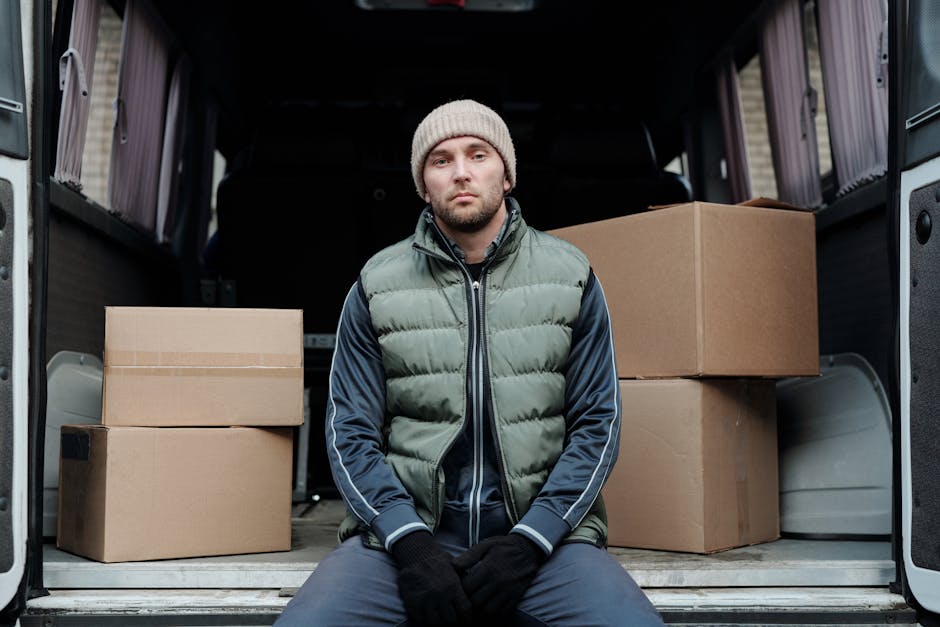 A man with a beard, wearing a blue beanie, red and navy jacket, black pants, and sneakers, stands with arms crossed in front of the open rear doors of a grey van loaded with multiple cardboard boxes d