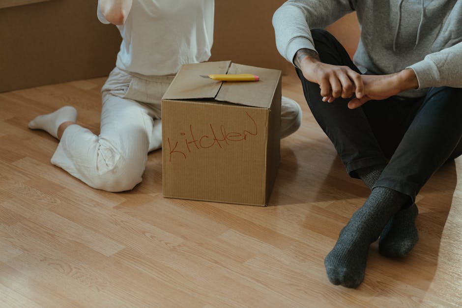 Two individuals are seated on a light wooden floor next to a cardboard box labeled 'kitchen' in red marker, surrounded by packing materials. One person, dressed in white trousers, is kneeling beside the box, holding a yellow marker on top of it, indicating an organized packing process typical of home relocation. The other person, wearing dark trousers and a grey sweater, is sitting cross-legged, with hands clasped, observing or assisting. Nearby, a partially visible door or doorway suggests they are inside a property preparing items for move. The scene reflects the packing and moving stage of house removals, with focus on proper labelling and handling of household items, and showcases the careful organization involved in furniture transport and the logistics of a residential move, as facilitated by [COMPANY_NAME], experts in removals.