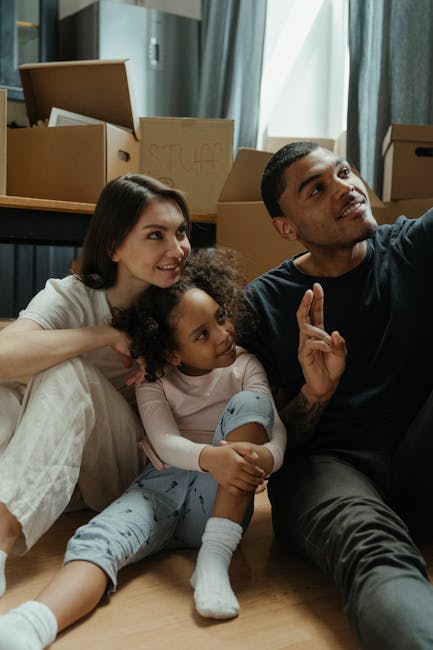 A family of three, including a woman with long brown hair, a young girl with curly hair, and a man with short dark hair, sitting on the floor inside a home surrounded by moving boxes made of cardboard and sealed with packing tape. The boxes, some labeled with words like 'STUFF,' are stacked nearby in a room with light-colored curtains and large windows allowing natural light to illuminate the space. The family appears relaxed as they sit close together, with the man gesturing with his hand, possibly explaining something, while the woman and girl listen attentively. The scene depicts a home relocation process, with unpacked boxes and an atmosphere of packing and moving, associated with domestic furniture transport and logistics handled by companies such as Man with Van Woolwich.